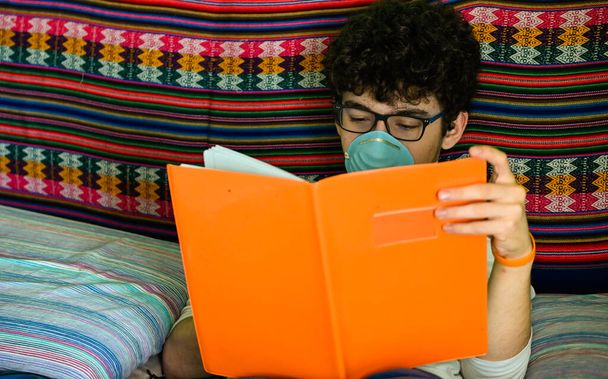 Un chico caucásico postrado en cama lleva una máscara protectora azul. Está estudiando su cuaderno cubierto de naranja. Concepto de estudio casero durante la cuarentena pandémica del coronavirus
. - Foto, Imagen