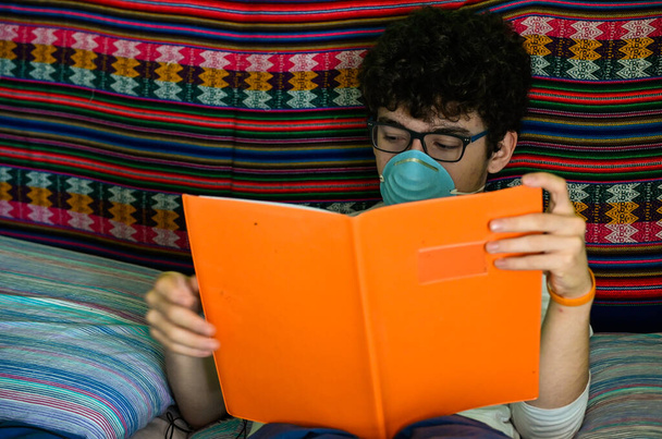 Un ragazzo caucasico costretto a letto indossa una maschera protettiva blu. Sta studiando il suo quaderno coperto di arancione. Home concetto di studio durante la quarantena pandemica coronavirus
. - Foto, immagini