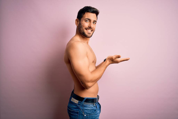 Jonge knappe sterke man met baard shirtloos staan over geïsoleerde roze achtergrond wijzen opzij met de handen open palmen tonen kopieerruimte, presenteren reclame glimlachend opgewonden blij - Foto, afbeelding