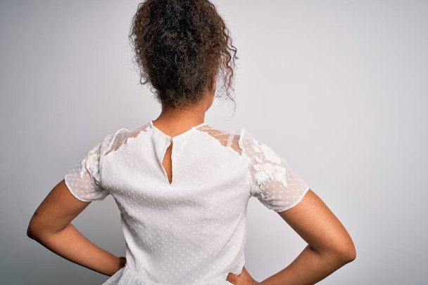 Jovem menina americana africana bonita vestindo t-shirt casual em pé sobre fundo branco de pé para trás olhando para longe com os braços no corpo
 - Foto, Imagem