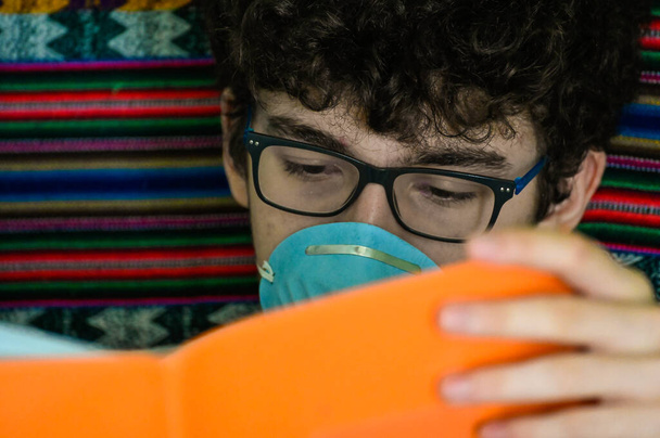 Πορτραίτο ενός κατάκοιτου καυκάσιου αγοριού με μπλε προστατευτική μάσκα. Μελετάει το πορτοκαλί τετράδιο του. Home study concept during coronavirus πανδημία καραντίνα. - Φωτογραφία, εικόνα