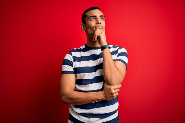 Handsome african american man wearing casual striped t-shirt standing over red background with hand on chin thinking about question, pensive expression. Smiling and thoughtful face. Doubt concept. - Fotó, kép