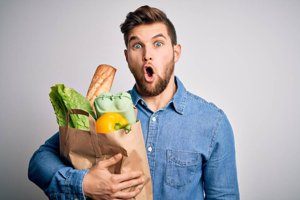Mladý blonďatý muž s vousy a modrýma očima držící papírový sáček s jídlem přes bílé pozadí vyděšený v šoku s překvapenou tváří, vyděšený a vzrušený výrazem strachu - Fotografie, Obrázek