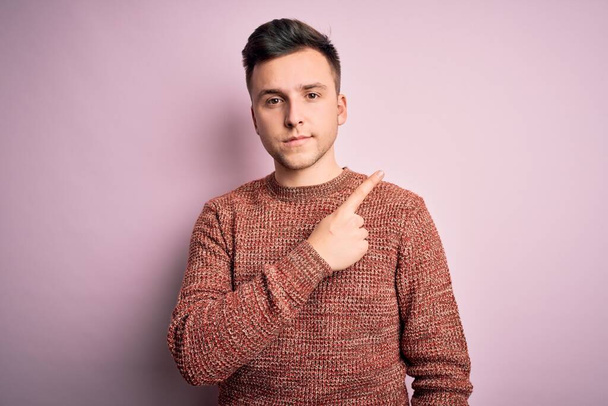 Joven hombre caucásico guapo usando suéter de invierno casual sobre fondo aislado rosa Señalando con el dedo de la mano al lado mostrando publicidad, cara seria y tranquila
 - Foto, Imagen