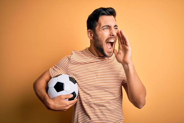 Όμορφος παίκτης με γενειάδα παίζει ποδόσφαιρο κρατώντας μπάλα ποδοσφαίρου πάνω από κίτρινο φόντο φωνάζοντας και ουρλιάζοντας δυνατά στο πλάι με το χέρι στο στόμα. Έννοια επικοινωνίας. - Φωτογραφία, εικόνα