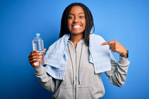 若いアフリカ系アメリカ人のフィットネス女性は、タオルを身に着けていると自分自身に指を指す驚きの顔を持つペットボトルから水を飲む - 写真・画像