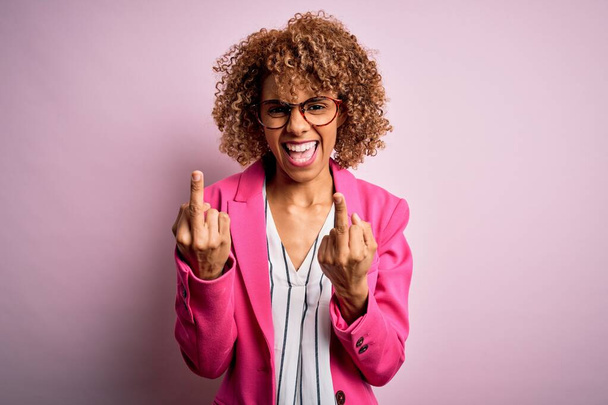 Молодая африканская деловая женщина в очках, стоящая на розовом фоне, показывает средний палец, делающий трахнуть вас плохое выражение лица, провокация и грубое отношение. Крики возбуждения
 - Фото, изображение