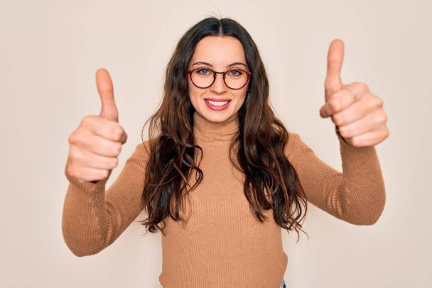 Young beautiful woman wearing casual turtleneck sweater and glasses over white background approving doing positive gesture with hand, thumbs up smiling and happy for success. Winner gesture. - Photo, Image