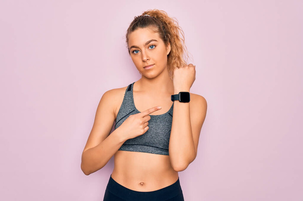 Young beautiful blonde sportswoman with blue eyes doing exercise wearing sportswear In hurry pointing to watch time, impatience, looking at the camera with relaxed expression - Photo, Image