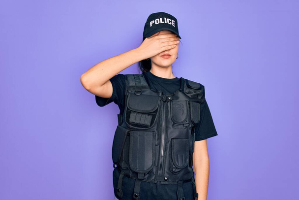 Egy fiatal rendőrnő golyóálló mellényt visel lila háttér felett, kezével befedi a szemét, komoly és szomorú arccal. Láthatatlan, rejtőzködő és elutasító koncepció - Fotó, kép