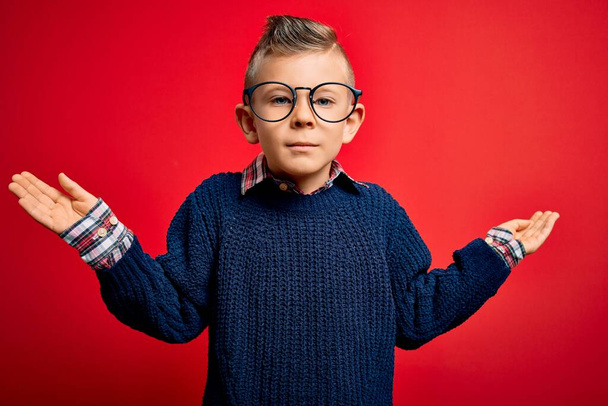 Ein kleines kaukasisches Kind mit blauen Augen steht mit einer intelligenten Brille vor rotem Hintergrund ratlos und verwirrt mit erhobenen Armen und Händen da. Zweifelhaftes Konzept. - Foto, Bild