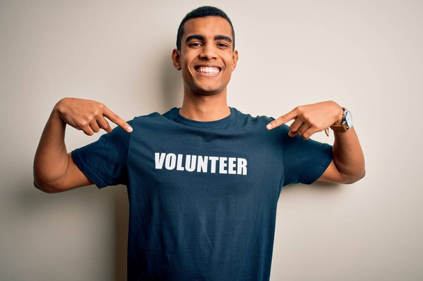 Mladý pohledný africký Američan, který se dobrovolně hlásí v tričku s dobrovolnickým vzkazem, vypadá sebevědomě s úsměvem na tváři, ukazuje prstem hrdě a šťastně. - Fotografie, Obrázek