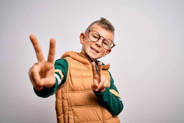 Ein kleines kaukasisches Kind mit blauen Augen, Wintermantel und smarter Brille, das lächelnd in die Kamera blickt und Finger zeigt, die ein Siegeszeichen machen. Nummer zwei. - Foto, Bild