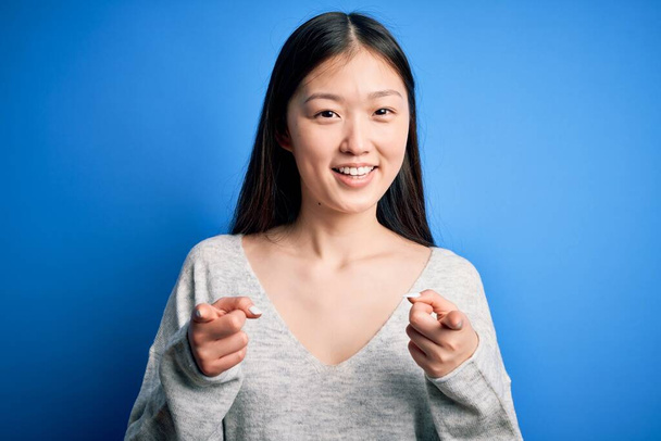 若い美しいアジアの女性が幸せと面白い顔を持つカメラに指を指して青い隔離された背景の上に立ってカジュアルなセーターを着て。良いエネルギーと振動. - 写真・画像