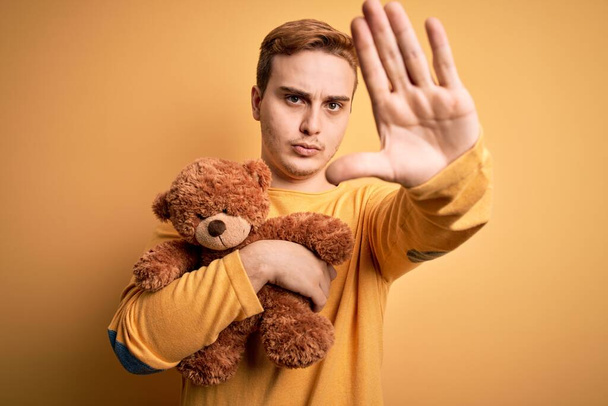 Jonge knappe roodharige man houdt teddy beer dier speelgoed over gele achtergrond met open hand doen stop teken met ernstige en zelfverzekerde uitdrukking, verdediging gebaar - Foto, afbeelding