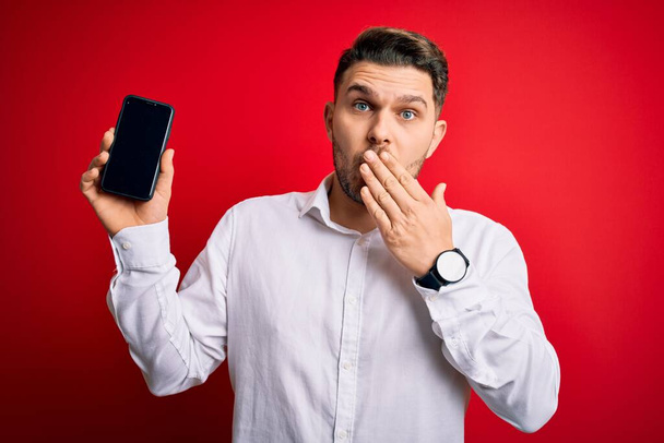 Młody biznesmen z niebieskimi oczami pokazujący ekran smartfona nad czerwonym tłem pokrywa usta z ręką zaszokowany wstydem za pomyłkę, wyrażaniem strachu, przerażony w ciszy, sekretna koncepcja - Zdjęcie, obraz