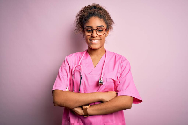 Afrykańska pielęgniarka w mundurze medycznym i stetoskopie na różowym tle szczęśliwa twarz uśmiechnięta skrzyżowanymi ramionami patrzącymi w kamerę. Osoba pozytywna. - Zdjęcie, obraz