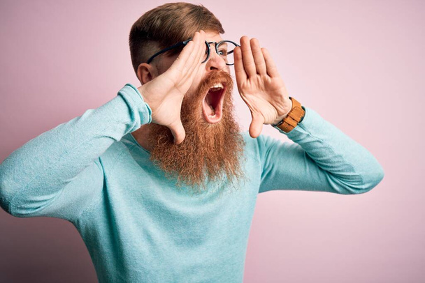 Knappe Ierse roodharige man met baard draagt bril over roze geïsoleerde achtergrond schreeuwen boos hardop met handen over de mond - Foto, afbeelding
