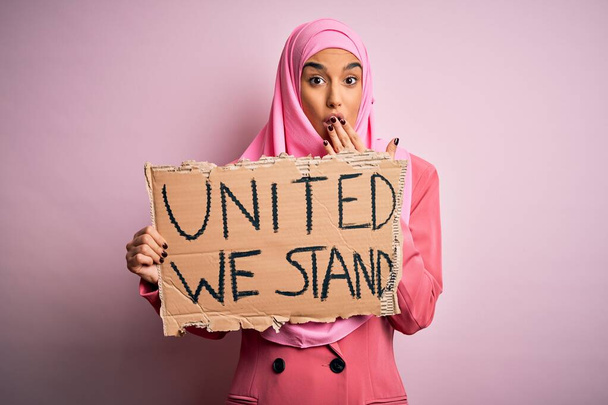 ピンクのイスラム教徒のヒジャーブを身に着けている若い活動家の女性私たちは、ミス、恐怖の表現、沈黙の中で怖がって、秘密の概念に恥でショックを受けた手で口をカバーメッセージをスタンド団結してバナーを保持 - 写真・画像