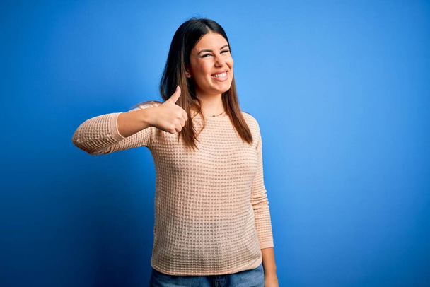 Young beautiful woman wearing casual sweater over blue background doing happy thumbs up gesture with hand. Approving expression looking at the camera showing success. - Photo, Image