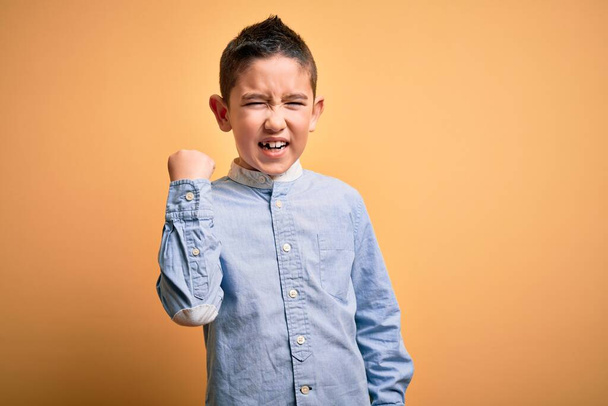 Νεαρό μικρό αγόρι φορώντας κομψό πουκάμισο στέκεται πάνω από κίτρινο απομονωμένο φόντο θυμωμένος και τρελός ανύψωση γροθιά απογοητευμένοι και έξαλλος, ενώ φωνάζει με θυμό. Οργή και επιθετική αντίληψη. - Φωτογραφία, εικόνα