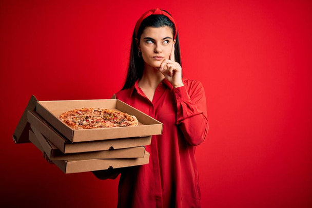 若いです美しいですブルネット女性保持配達ボックスとともにイタリアのピザ以上赤の背景深刻な顔考える上で質問,非常に混乱したアイデア - 写真・画像