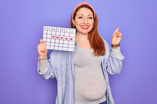 Junge schöne rothaarige schwangere Frau erwartet Baby Wartezeit Kalender sehr glücklich zeigt mit der Hand und dem Finger zur Seite - Foto, Bild