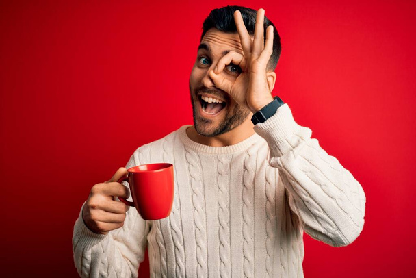 Genç yakışıklı adam kırmızı izole edilmiş arka planda bir fincan sıcak kahve içiyor. Gülümsüyor. Tamam işaret veriyor. - Fotoğraf, Görsel