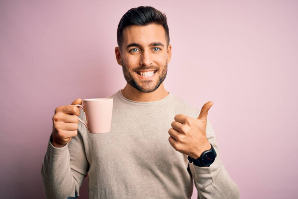 Nuori komea mies juo kupin kuumaa kahvia vaaleanpunainen eristetty tausta onnellinen iso hymy tekee ok merkki, peukalo ylös sormilla, erinomainen merkki
 - Valokuva, kuva