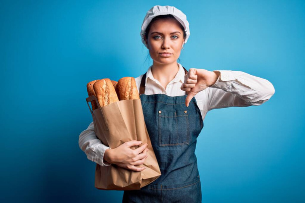 Mladá krásná pekařka s modrýma očima držící papírový sáček s chlebem s rozhněvaným obličejem, negativní nápis ukazující odpor s palci dolů, koncept odmítnutí - Fotografie, Obrázek