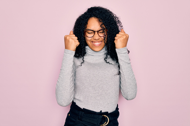 Young african american woman wearing turtleneck sweater and glasses over pink background excited for success with arms raised and eyes closed celebrating victory smiling. Winner concept. - Фото, изображение