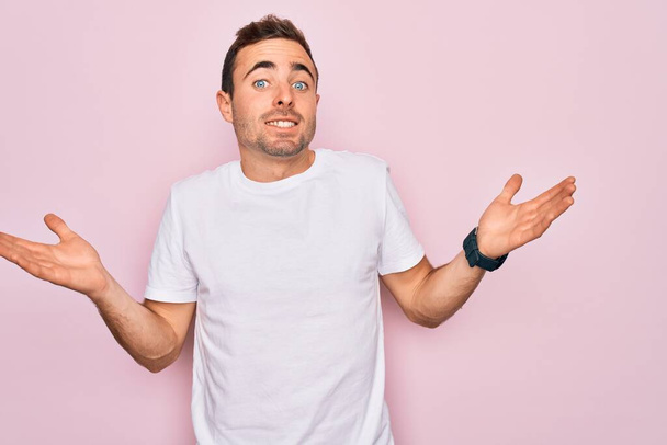Hombre guapo con ojos azules que usa una camiseta blanca casual de pie sobre un fondo rosado sin pistas y expresión confusa con los brazos y las manos levantadas. Concepto de duda
. - Foto, Imagen
