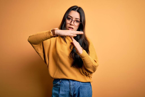 Mladá brunetka žena nosí brýle a ležérní svetr přes žluté izolované pozadí dělá time out gesto s rukama, frustrovaný a vážný obličej - Fotografie, Obrázek