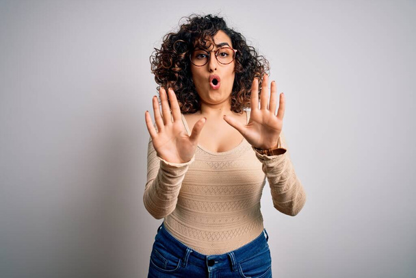 Junge schöne, lockige arabische Frau mit lässigem T-Shirt und Brille vor weißem Hintergrund, die ihre Hände mit ängstlichem und ekelhaftem Gesichtsausdruck von den Handflächen wegbewegt und Verweigerung und Verweigerung zeigt. Stopp und Verbot. - Foto, Bild