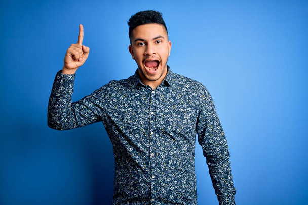 Ein junger gutaussehender Mann in lässigem Hemd steht vor isoliertem blauen Hintergrund und zeigt mit erhobenem Zeigefinger auf eine erfolgreiche Idee. Aufgeregt und glücklich. Nummer eins. - Foto, Bild