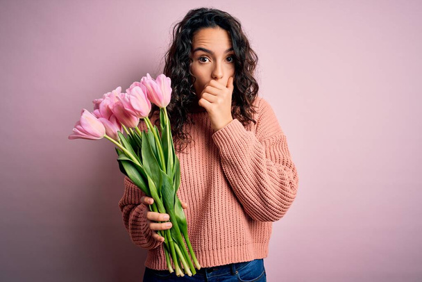 Young beautiful romantic woman with curly hair holding bouquet of pink tulips shocked covering mouth with hands for mistake. Secret concept. - 写真・画像