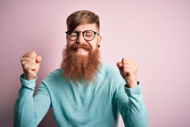 Komea irlantilainen punapää, jolla on parta silmälasit vaaleanpunaisen eristetyn taustan päällä, innoissaan menestyksestä kädet kohotettuina ja silmät suljettuina juhlimassa voittoa hymyillen. Voittajan käsite
. - Valokuva, kuva
