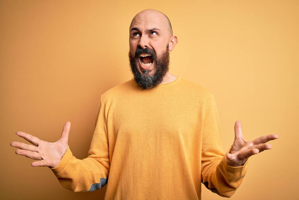 Handsome bald man with beard wearing casual sweater standing over yellow background crazy and mad shouting and yelling with aggressive expression and arms raised. Frustration concept. - Foto, Bild