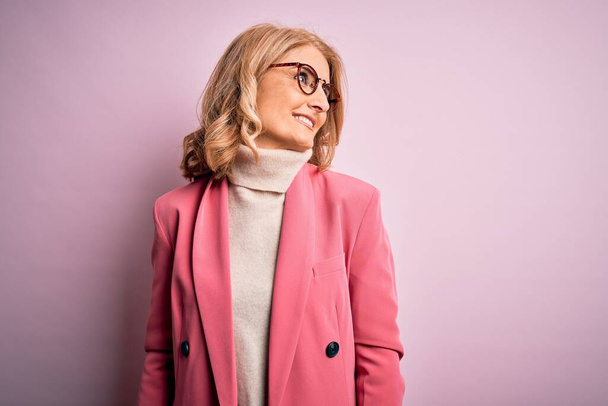 Mulher de negócios loira de meia idade bonita vestindo jaqueta rosa elegante e óculos olhando para o lado com sorriso no rosto, expressão natural. Rindo confiante
. - Foto, Imagem