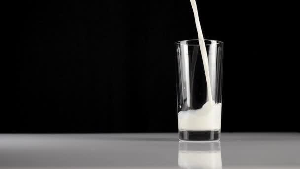 Čerstvé bílé mléko se nalije do průhledné sklenice na bílém stole na černém pozadí. Koncept zůstat doma a jíst zdravé jídlo. Výborná snídaně. Široký záběr. Zpomalený pohyb - Záběry, video