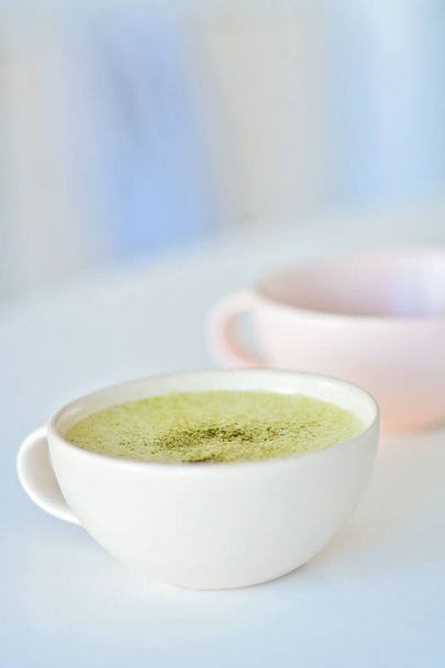 White cup with matcha latte on a white table, a light pink cup on the background, Matcha green tea latte in a cup. Top view. Copy space. Green tea matcha latte is a delicious way to enjoy the energy boost and healthy benefits of matcha. Matcha is a p - Photo, Image