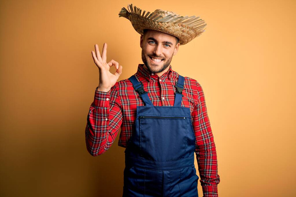 Young rural farmer man wearing bib overall and countryside hat over yellow background smiling positive doing ok sign with hand and fingers. Successful expression. - Foto, immagini