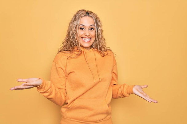 Jonge mooie blonde sportieve vrouw dragen casual sweatshirt over gele achtergrond glimlachend vrolijk met open armen als vriendelijke ontvangst, positieve en zelfverzekerde groeten - Foto, afbeelding