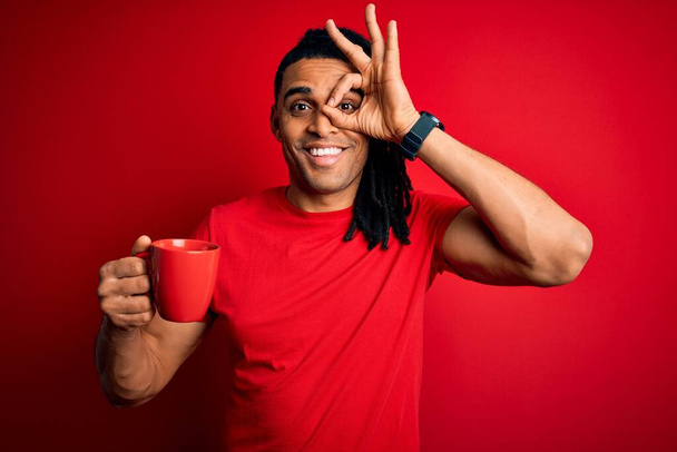 Joven afroamericano afro hombre bebiendo taza de café sobre fondo rojo con la cara feliz sonriendo haciendo signo bien con la mano en el ojo mirando a través de los dedos
 - Foto, imagen