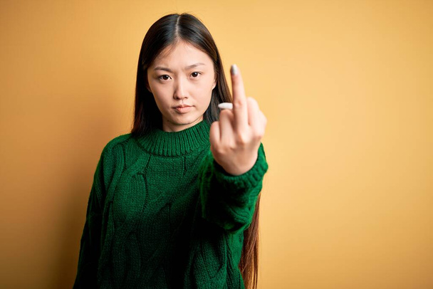 Joven hermosa mujer asiática vistiendo verde invierno suéter sobre amarillo aislado fondo Mostrando el dedo medio, descortés y grosero joder off expresión
 - Foto, imagen
