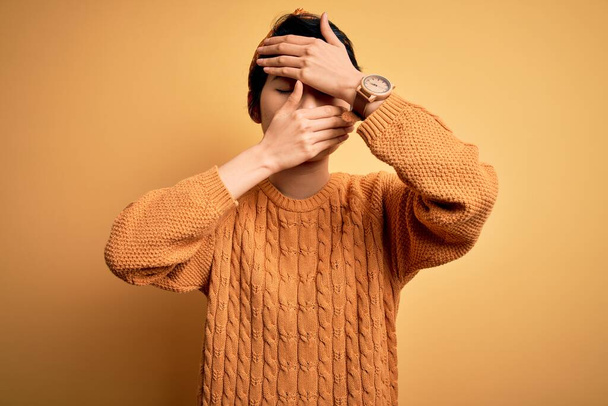 Young beautiful asian girl wearing casual sweater and diadem standing over yellow background Covering eyes and mouth with hands, surprised and shocked. Hiding emotion - Photo, Image