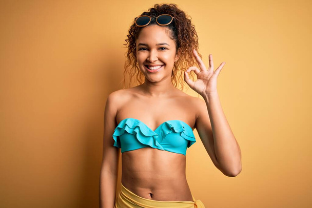 Junge schöne Amerikanerin im Urlaub im Bikini vor isoliertem gelben Hintergrund, die positiv lächelnd ein Okay-Zeichen mit Händen und Fingern macht. Erfolgreicher Ausdruck. - Foto, Bild