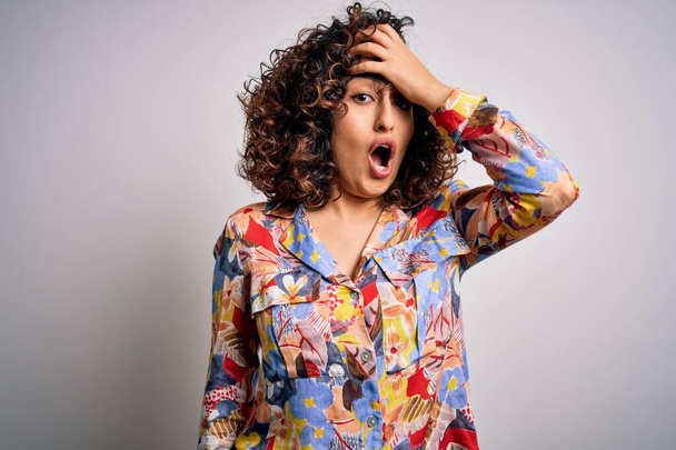 Young beautiful curly arab woman wearing floral colorful shirt standing over white background surprised with hand on head for mistake, remember error. Forgot, bad memory concept. - Photo, Image