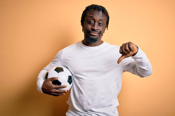 Giovane giocatore afroamericano uomo che gioca a calcio tenendo palla da calcio su sfondo giallo con la faccia arrabbiata, segno negativo che mostra antipatia con i pollici verso il basso, concetto di rifiuto
 - Foto, immagini