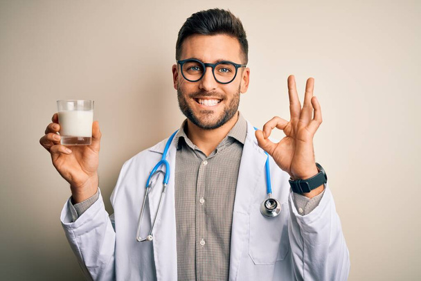 Young doctor man wearing stethoscope holding a glass of milk over isolated background doing ok sign with fingers, excellent symbol - Fotografie, Obrázek